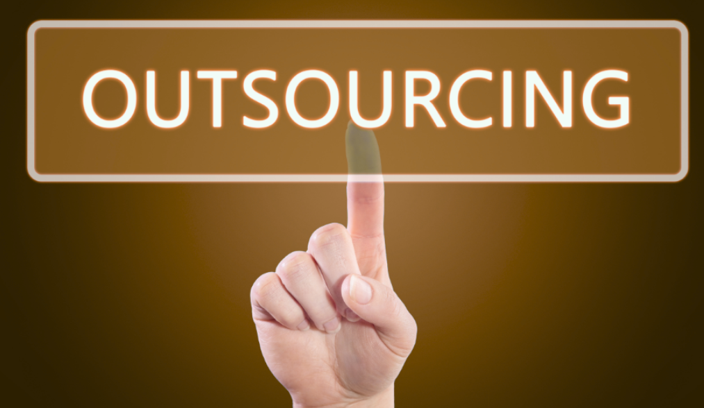 Hand zeigt auf das Wort "OUTSOURCING" vor einem braunen Hintergrund.